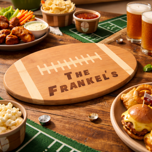 Wooden football-shaped cutting board with 'The Frankel's' engraving on a table with snacks and drinks.